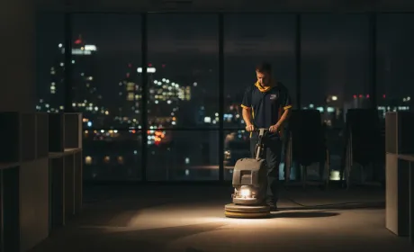 An Australian commercial cleaner in branded polo and hi-vis buffing the floor of an empty open-plan office at night, CBD lights through the glass behind them