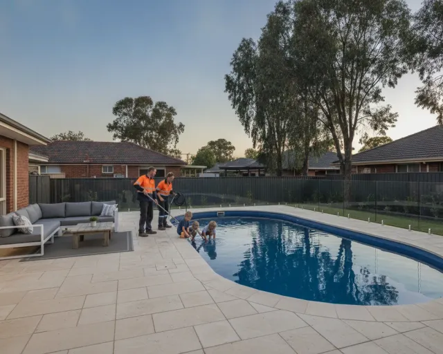 A finished fibreglass pool at dusk in a Perth backyard, frameless glass pool fence, limestone coping and paving, outdoor lounge setting and a family beside the water
